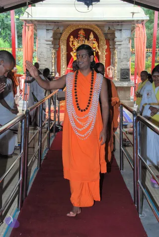 A barefoot man in saffron-orange robes walks down a red carpet between metal railings toward a temple shrine, raising his right hand in blessing. He wears a dark rudraksha mala and several layers of white beaded garlands over his shoulders, with a red bindi on his forehead. Behind him, a monk in orange follows, and devotees in white with yellow shawls stand on either side with hands folded. The shrine behind has a golden arch, orange draped curtains, and a gilded deity murti adorned with garlands. A camera tripod and cameraman are visible to the left. The shoulder-length swept-back hair, saffron robes, rudraksha mala and ceremonial garlands match the page's Swamiji claim, so this is a probable match.