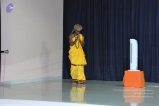 A photograph of a single standing figure on a polished stage in front of a dark navy curtain. The person wears a yellow saffron-style dhoti with a gold-toned border, a tan turban, and a dark rudraksha-style bead mala. Palms are pressed together in front of the chest in a namaskar greeting, with head slightly bowed. A white tower fan on an orange-draped block stands at right, and a microphone stand is visible at left.