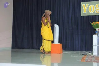 A standing man on a stage faces left with palms pressed together in a namaste greeting. He wears a tan turban, a yellow silk garment with a gold border draped over bare shoulders, and a dark rudraksha-style bead mala around his neck. Behind him a dark navy curtain hangs, a green banner with yellow letters reading "Yog" is at upper right, and a vase of orange and yellow flowers sits at the right edge beside a white cabinet. A white tower fan stands on an orange-draped box near him, and the polished floor reflects the figure. a person: the figure's long-format presentation includes a turban, bead mala and a yellow ceremonial garment, presented as a satsang speaker.