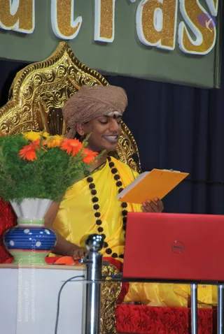 A photograph of a man seated on an ornate gold throne-style chair on a stage, dressed in a yellow silk garment and wearing a pale brown turban with a red bindi and tilak on the forehead. He wears a long string of dark rudraksha beads and holds an orange clipboard while smiling and reading. In front of him are a vase of orange and yellow flowers with greenery and a red laptop on a stand. Behind him is a green banner with large gold lettering partly visible at the top, and dark blue curtains.