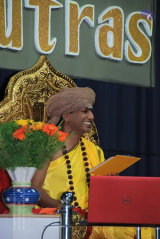 A smiling man in a brown turban and yellow silk garment sits before an ornate gold throne-style chair, holding an orange clipboard and wearing a long rudraksha mala. A red laptop sits on a metal stand in the foreground, and a vase of orange and yellow flowers with green foliage stands at left. Behind him, large gold glitter letters on a green banner read 'utras' (part of 'Yoga Sutras'), with a dark blue curtain behind. A white tilak marks his forehead.
