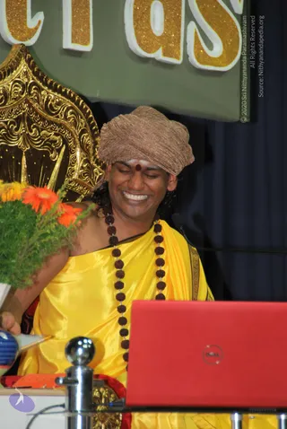 A smiling man with closed eyes sits on an ornate gold throne-style chair at a stage talk. He wears a bronze-gold turban, a red bindi and white forehead markings, a saffron-yellow silk garment and a long rudraksha mala. A red laptop sits in front of him, a microphone stands nearby, and a flower arrangement with orange gerberas is at left. Behind him is a green banner with gold glitter lettering and dark blue drapes. Probable Swamiji by visible cues of ornate clothing, turban and rudraksha mala, seated in his role as speaker at the satsang.
