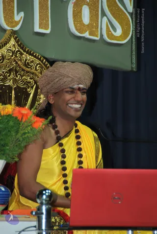 A man seated on an ornate gold throne laughs broadly beside a red Dell laptop, with orange gerbera flowers and green foliage at the left edge. He wears a brown metallic turban, a yellow silk garment with a decorative border, and a long rudraksha mala with a vertical tilak and red bindi. Behind him a green banner reads partly in gold glitter lettering, with dark blue drapery and a stage microphone stand in view. Appearance-based label: Swamiji (probable), seated in a satsang setting, with long hair and rudraksha malas visible.