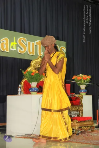 A barefoot man in a yellow silk garment with gold border stands on a glossy stage, palms pressed together in anjali namaskar with head bowed. He wears a tan turban, a dark rudraksha mala, and his hair falls past his shoulders. Behind him is a green banner reading Yoga Sutras, a gold-decorated chair with red cushion, a white table holding flower vases with orange gerbera and greenery, and a golden ornament. Dark blue curtains form the backdrop. Swamiji (probable) appears to be the figure, based on the yellow saffron-style clothing, long hair, turban and rudraksha mala. He is the only person visible.