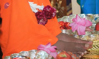 Close-up photograph of a pada puja, the foot-worship ritual. Bare feet rest on a golden tray surrounded by silver bowls holding red and pink flowers and sliced fruit. A pink lotus blossom lies on the feet and another on the tray. The person wears an orange saffron robe with a garland of white and magenta flowers draped over the shoulder. A second person in blue clothing appears at the upper right edge.