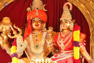 Golden murti images of a deity couple seated side by side against a deep red draped backdrop framed by an ornate gold arch. The male figure on the left wears an orange-red striped turban with a crescent moon, a gold trident (trishula) in his raised hand, and layered gold and jewelled necklaces with green and red gemstones. The female figure on the right wears a red sari with gold zari border, a tall jewelled crown, and heavy gold necklaces. Both are garlanded with yellow and purple flowers, and small oil lamps glow at the base in front. These are statues, not living people.