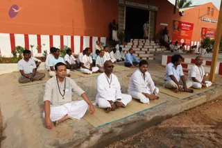 A photograph of a group of people seated in meditation on woven mats on a paved courtyard in front of a red and white painted temple facade. A sign above the doorway reads Sri Anandeshwara Temple. Most men wear white dhotis and shirts, some in grey or light blue, with rudraksha malas around their necks. Several sit with eyes closed, hands resting on their knees. A red and white striped border runs along the wall and stairs, and a banner on the right advertises a Life Bliss Program. A statue stands at the left side of the entrance. Approximately twenty people are visible.