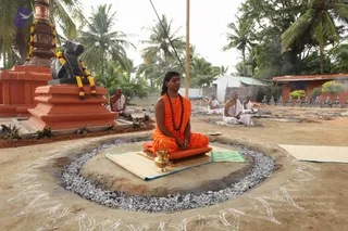 Outdoor photo of a seated man in saffron-orange robes with a rudraksha mala and a red-orange forehead mark, meditating cross-legged on a mat over a circular raised earth platform ringed with grey gravel, with a brass vessel beside him. Behind him, several men in white dhotis and shawls sit in meditation on mats across the sandy ground, near a black stone Nandi bull decorated with orange and yellow marigold garlands on an orange pedestal, palm trees and a thatched wall behind. Chalk-drawn kolam patterns mark the ground. A row of small grey figurines stands along a wall at right.