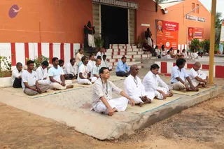 A group of about twenty people sit cross-legged in meditation on mats outside a terracotta-coloured temple with red and white striped borders. The temple sign reads Sri Anandeshwara Temple. Several men in white dhotis and kurtas, and others in light shirts, sit in rows on the paved platform in front of steps where more people sit. Two stone statues in saffron-draped robes stand flanking the doorway. A red banner on the right advertises a Life Bliss Program with a 2 days meditation camp. A man in the front centre wears a light grey kurta and a rudraksha-style necklace, and a bald man in a white kurta sits beside him wearing dark glasses and a beaded necklace.