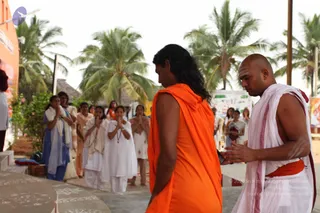 Outdoors under palm trees, a man with long wavy dark hair wearing an orange saffron wrap stands with his back to the camera and head lowered, facing a shaven-headed priest in white ritual cloth with a red border and a red waist sash. The priest holds his palms together in front of him. Behind them, a row of women in white and pale clothing stand with palms pressed together in namaste, and a thatched hut and a white banner with green and blue print are visible.