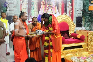 A ceremonial scene in a decorated hall with a painted temple backdrop of gopuram-style architecture. In the centre, a long-haired figure in orange robes wears a heavy garland of green leaves, yellow and red flowers and stands before a gilded throne with red cushions. He bows with a hand on his forehead. To the left, a man with a shaved head and white tripundra markings, wearing an orange dhoti and a rudraksha-style orange bead necklace, holds a silver tray of bananas and offerings toward him. Beside him, a shaved-headed attendant in orange robes looks down. Behind them, several men stand, including one in a white dhoti and yellow shawl. Brass trays with flowers and small oil lamps sit in the lower right foreground. The image appears to show a padapuja-style offering to a seated guru. a person candidate by appearance: long hair, saffron garments and a garland on the person at the throne.