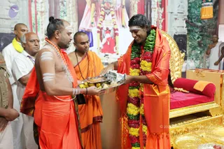 A photograph of a ceremonial moment in a decorated hall. On the right, a smiling man with long swept-back dark hair wears an orange-saffron shawl and a large garland of red, yellow and green flowers, seated before a gold-and-red throne with a red cushion. On the left, a bare-chested priest in orange cloth with white ash markings on his torso and forehead holds a silver tray of fruit and offerings toward him. Beside them stands an attendant in orange robes with a rudraksha mala, and further left a man in white with a yellow shawl. A backdrop shows a deity image with flower garlands.