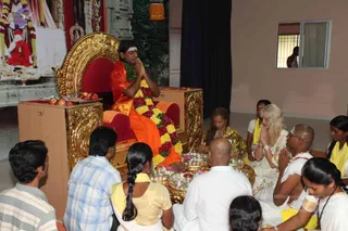 A seated man on an ornate gold-and-red throne with his palms pressed together in namaste, wearing an orange-saffron shawl and a long green-and-red flower garland, with a white tilak on his forehead. Before him on a low gold table are brass plates of offerings, flowers and fruit, with a red-cloth-covered stool and a fruit bowl nearby. Around him sit devotees on the floor: a bald man in white dhoti with a sacred thread and gold necklace, a woman in a yellow sari, a blonde woman in white with folded hands, a woman in a gold-patterned top, and several people in yellow and white clothes, plus a person in a striped shirt at the left edge. Behind is a shrine backdrop with a red-robed deity image and garlands. This appears to be a pada puja ritual with devotees honouring the seated figure.