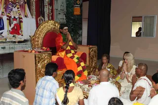 A photograph of a padapuja-style devotional gathering in an indoor hall. A man seated on an ornate gold-and-red throne chair, wearing an orange-saffron shawl and a long multicolour garland of marigolds and red roses, holds his palms together in namaskar, with a vertical white tilak on his forehead. Devotees sit on the floor in front of the throne, including a man in a striped shirt, a woman in a yellow sari with a long braid, a bald man in white ritual clothes, and women in yellow and white saris with hands folded in prayer. Silver trays holding brass plates and small lamps sit near the throne, along with a fruit tray with red apples on the left side. Behind the throne a large backdrop shows a painted temple image with a red-clad figure. A partly visible man looks through a side window on the right.