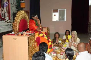 A seated man in an orange-red garment with a green and red flower garland stands at a gilded throne with a red cushion, hands pressed together in namaste and a white tilak on his forehead. Before him, devotees sit on the floor behind gold-rimmed brass trays holding flowers, fruit and small oil lamps, part of a pada puja offering. A woman in gold sari with a name badge and a woman in yellow with folded hands kneel nearby, while a woman in a white and gold outfit and a bald man in white join in prayer at the lower right. Apples and flowers sit on a gilded table behind the throne, with a shrine and decorated pillars at the left edge.