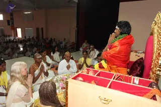 A photo from a hall where a man with long dark hair, wearing an orange-red shawl and a green-and-white flower garland, stands with palms pressed together in namaskar on a raised stage beside a gilded throne with a pink cushion. In front of him a wooden offering box with red compartments holds apples, and beside it sits a silver tray with pink flowers and a small oil lamp. Seated devotees fill the floor: a bald man in white with a tilak and beaded necklace, a woman in white with folded hands, a woman in a yellow sari, and a woman with braided hair facing away in the foreground. Behind them a large audience in white clothes sits in rows. Swamiji (probable) is the man in orange-red on the stage, identified by his long swept-back hair, orange-red shawl, garland and position on the throne-side dais.