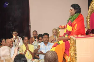A photograph of a pada puja during a satsang. On the right, a man with long swept-back dark hair, a white forehead mark and a saffron-orange shawl over a green and red flower garland sits on a gold-trimmed red throne, eyes closed with hands extended forward. A probable Swamiji candidate with long hair, saffron clothing and a garland. Before him, a low wooden stand holds fruit and a brass tray. Seated devotees in the foreground include a woman in a yellow sari with palms pressed together, a man in a light blue checked shirt holding a small oil lamp, and a bald priest in white holding a lit lamp. Behind them, shaved-headed priests in white dhotis and beaded necklaces sit, one holding a microphone. Foreground heads of audience members are visible at the bottom.