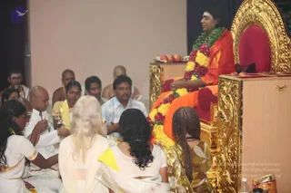A photo of a seated discourse in a hall. On a raised gold-carved throne at upper right sits a figure in saffron-orange clothing with a long green and red-and-yellow flower garland, a red cushion behind him, and a fruit offering on a tray beside him. He appears to be speaking. Seated before him on the floor are devotees: a bald man in white holding an oil lamp at left, a man in a white shirt and a woman in yellow near the centre, a woman in a white sari with blonde hair in the foreground, a woman with dark wavy hair in a white sari with a yellow border, and a woman with braided hair in a floral dress. A microphone stands at upper left, and a gold-carved lectern is at right.