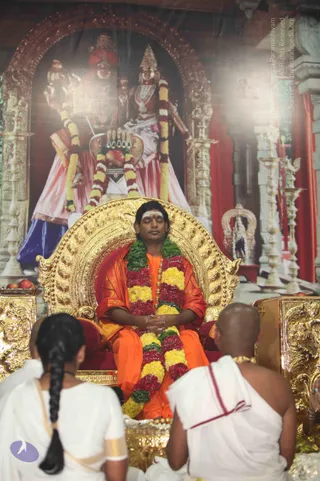 A photograph of a man seated on an ornate gold throne with eyes closed and hands folded, wearing an orange saffron robe, a multicoloured flower garland of red, yellow and green blooms, a white tilak on his forehead and a red dot. Two people in white ritual clothing sit with their backs to the camera in the foreground: a woman with a long black braid and a shaved-headed man in a white dhoti with a red border. Behind the throne is a large painted backdrop of two standing deities in red and pink garments with tall crowns, flanked by brass lamp stands and red curtains.