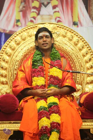 A seated man speaks into a microphone on an ornate gold throne with a carved arched back. He wears an orange saffron robe with an orange-red silk shawl, a long garland of yellow and red flowers with green leaves draped across his chest, and a white vibhuti marking with a red kumkum dot on his forehead. His hands are folded in his lap. Behind him a large pink and white draped backdrop displays a framed portrait garlanded with flowers. Red cushions flank the throne. Swamiji (probable): long swept-back hair, saffron robes, flower garland and ornate gold throne, presiding as a speaker at a satsang talk.