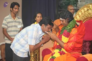 A photograph of a seated figure on an ornate golden throne with a red cushion, wearing an orange-red silk garment and a long garland of yellow and magenta flowers with green leaves. The figure has long swept-back dark hair, a white tilak on the forehead, and a raised hand touching the head of a man in a light blue and white checked shirt who bends forward to bow. Behind them a standing woman in a green-and-white setting wears a red bindi, another man in orange stands at the right, and a young man in a striped grey shirt stands at the left. Dark blue drapery forms the backdrop. Orange-clad figure with long hair and distinctive adornments, seated on the throne, receiving the bow of a devotee.