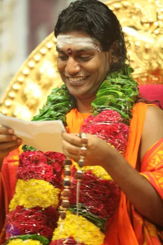 A smiling man with long, swept-back dark hair and a white horizontal tilak with a red dot on his forehead sits before a gold ornate throne. He wears an orange-saffron garment and a green leaf garland layered over red and yellow flower garlands, and holds a sheet of paper while a string of rudraksha beads hangs from his hand. Swamiji (probable): the long swept-back hair, saffron garments and rudraksha malas match the page's identification of the Supreme Pontiff at a satsang. One physical person is visible.