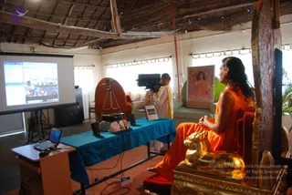 A seated man with long swept-back dark hair wears a saffron-orange robe and sits on an ornate gilded throne carved with a lion-like figure, in a bright hall with a thatched ceiling. A cameraman in white with a yellow shawl films him with a camera on a tripod. A projection screen on the left shows a video of a crowd and a lectern-style laptop sits nearby. A framed painting of a seated woman in a red-orange tone hangs on the wall behind, and a blue tablecloth covers a long table. Swamiji (probable): the seated man wears a saffron robe, long swept-back hair and sits on a gilded throne, a presentation cue consistent with a speaker at a satsang.