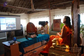 A seated man in an orange saffron robe with long swept-back hair sits on an ornate carved wooden throne with gold lion-head carvings, beside a blue draped table. He is the probable Swamiji, identified by long hair and saffron clothing, and is in the room where a live talk is being filmed. Behind the table, a cameraman in white and yellow clothing films with a video camera on a tripod. A projector screen at left shows a video call with participants. A framed portrait of a woman in orange robes hangs on the wall, and a thatched roof overhead.