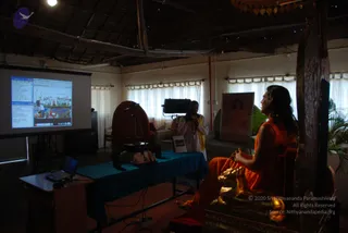 A photograph of a meditation hall with a thatched ceiling and white curtains. On the right, a man with long dark hair wears an orange robe and sits on a carved wooden throne with gold lion-head armrests, hands folded, facing left. A second man in white clothing with a yellow shawl stands near a camera on a tripod, a small figure of the head on a blue-draped table. A projector screen on the left shows a web page with a group photograph of people outdoors. A framed portrait of a woman in orange hangs on the back wall beside a potted plant. Probable Swamiji seated on the throne in orange robes with long hair, with a gold lion-headed armrest and a tall wooden backrest.