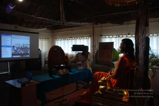 A photograph of a dimly lit ashram hall with a thatched wooden ceiling and white-curtained windows. On the right, a man with long dark hair wearing an orange robe sits on an ornate gilded chair with a carved wooden frame, facing left toward a projection screen. A cameraman in white clothing with a yellow shawl stands behind a tripod camera near a teal-draped table. The projected screen shows a web page with a group photograph of people. A framed portrait of a woman in orange-pink robes hangs on the wall, and a potted palm sits beside the chair. A laptop rests on a small wooden desk at left. The man's figure is partly silhouetted, and his face is not clearly visible.