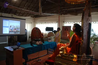 A photo of a live satsang in a thatched-roof hall with white walls and curtained windows. A long-haired man in orange robes sits on an ornate carved wooden throne with gold lion-shaped armrests, hands raised in a gesture while speaking. Behind him a cameraman in white and a yellow shawl films with a tripod-mounted camera. A projector screen on the left shows a web page with text and photos, and a laptop sits on a wooden stand. A blue draped table holds equipment, and a framed painting of a woman in pink hangs on the wall. The man is an appearance-based a person, distinguished by long swept hair and saffron robes on a throne, so this is an in-person candidate.