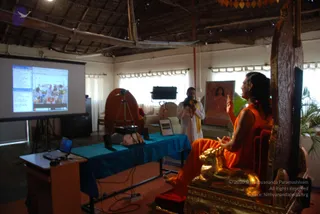 A photograph of a satsang talk in a thatched-roof hall with white walls and cream curtains. A seated man in orange robes with long swept-back dark hair sits on an ornate golden-carved wooden throne, gesturing with raised hands while speaking. He has a rudraksha-style beaded mala and appears to fit the appearance cues associated with a person. Behind him stands a man in white clothes with a yellow shawl holding a microphone. A projector screen on the left shows a webpage and video feed. A blue-draped table holds a laptop and equipment, and a framed portrait of a woman in pink hangs on the wall. Plants and a tripod with a lamp complete the setting.