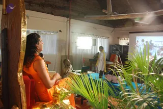 Indoor satsang room with a thatched ceiling and white curtained windows. A long-haired figure in saffron-orange robes and a gold-embroidered shawl sits on a red-cushioned seat with a carved wooden backrest, facing a projection screen on the right. Two other people in white are visible: one seated at a table with a laptop, and another standing near a tripod-mounted camera beside a blue-draped table. A potted palm sits in the foreground. The projected screen shows a group of people. A gold-framed backrest and a visible tripod suggest a recorded talk.