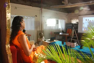 A seated person in an orange sleeveless robe with long dark hair sits at left on a red chair, facing a projector screen, hands clasped in the lap. Beside them, another person in orange and a man in white dhoti and shawl sit in a plain room with white curtains and a blue-draped table holding a laptop and a projector on a tripod. The screen shows a photo of a figure in white with a crowd. Palm fronds frame the foreground and a gold-edged wooden panel sits at left. a person candidate is present, identified by long swept hair and orange robes; the appearance cues are reasonably distinct.
