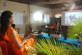 A indoor room with a white wall, exposed wooden ceiling beams and white curtains at the windows. In the foreground on the left, a man with long dark swept-back hair wears an orange-saffron garment, a white-and-gold garland and a gold-coloured fabric draped over his lap, seated in a red chair and looking toward a projector screen. Beside him sits another man in an orange-saffron garment and a garland. In the centre, a shaved-headed man in white dhoti and white garment sits at a table with a laptop. Green palm fronds frame the foreground. A projector screen on the right shows a photograph of a crowd of people, and a tripod-mounted camera stands on a blue-draped table.