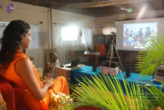A woman with long dark wavy hair in an orange sleeveless top sits in a red armchair at left, hands clasped in her lap, gazing toward a projection screen. Beside her, a bald man in white ritual clothing with a beaded necklace sits at a wooden desk with a laptop. A projector on a tripod and a blue-draped table are in the foreground, with a palm frond at right. The screen shows a crowd of people. Strong studio lighting illuminates the plain white-walled room. The orange-clad person is a devotee, not clearly identifiable as a person from appearance alone, so the identity remains uncertain.