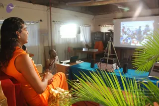 A photo of a bright indoor room during a talk session. In the left foreground a long-haired man in an orange sleeveless garment with a flower garland sits on a red chair with clasped hands, looking toward a projection screen on the right showing a photograph of a crowd. Behind him a man in white dhoti and shawl sits at a wooden desk with a laptop, and a blue-draped table holds a projector on a tripod and speakers. Green palm fronds frame the foreground, a golden object sits at lower left, and white curtains and a window light the room.
