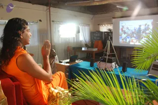 A photograph of a long-haired man in a sleeveless orange robe seated on a red cushioned chair at the left edge, hands joined in front of his chest in an anjali gesture while he faces right toward a projection screen. A second, shaved-headed man in white sits behind a wooden desk with a laptop in the background. A tripod-mounted camera, speakers, and a blue-draped table sit in the foreground, with palm fronds in front of the lens. The projected slide shows a group of people gathered around tables. The man in orange is an appearance-based probable Swamiji, identifiable by his long swept-back hair and orange clothing, seated during what the event listing describes as a satsang talk. The room has white curtains, a bookshelf, and an exposed ceiling.