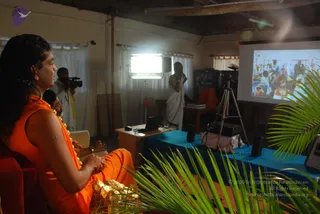 A photograph of an indoor satsang room with a projection screen showing an image of people gathered. In the foreground at left, a man with long dark swept-back hair wears an orange saffron robe with a garland of flowers and sits with hands clasped, looking toward the screen; a visible ceremonial gold necklace and rudraksha-style adornment are present. Beside him, another seated man in orange partly visible. A standing man in a white dhoti and shawl speaks into a microphone near a table with a blue cloth, a laptop and speakers. A cameraman with a video camera stands at the back left beside a bright light panel. Green palm fronds frame the foreground.