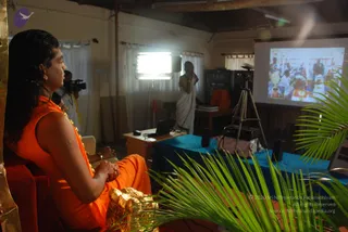 A photograph of an indoor satsang room with a projector screen at right showing a crowd photo. In the foreground at left, a person with long swept-back dark hair wearing an orange sleeveless garment and a garland sits on a red chair with hands folded, facing the screen; the appearance is consistent with a person based on long hair and saffron clothing. A standing man in white dhoti and shirt speaks beside a blue-draped table with a laptop and speakers. A cameraman with a camera stands behind at left. Palm fronds appear in the foreground and a tripod and light stand are visible.