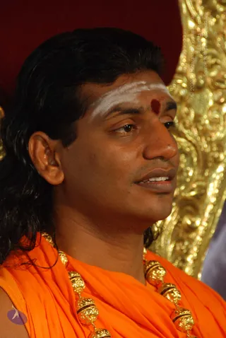 Close-up photograph of a man in profile wearing an orange saffron shawl draped over the shoulders, with a heavy gold necklace of ornate beads. He has long swept-back dark hair, a white vertical tilak on the forehead and a red kumkum dot between the brows. A golden ornate throne or decorative backdrop is visible behind him against a red fabric. Probable Swamiji: long swept-back hair, saffron clothing and ornate gold necklace are present, with the figure appearing to be seated in a ceremonial setting.