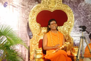 A man with long swept-back dark hair sits smiling on an ornate gilded throne with a red velvet backrest and golden lion-head armrests. He wears a saffron-orange sleeveless robe, a long strand of large gold-coloured beads, and a white and red tilak on his forehead, and holds a bundle of gold bangles in his hands. Behind him is a backdrop of trees, and a camera on a tripod is visible at right beside a partly visible person in orange.