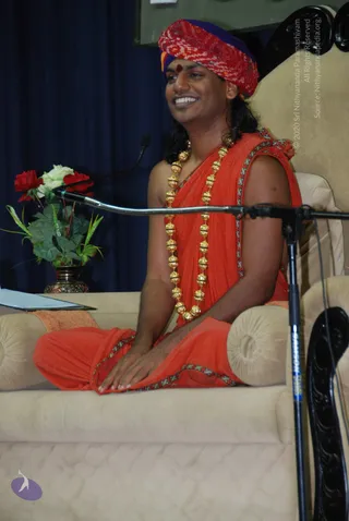 A man seated cross-legged on a cream upholstered throne-like seat before a microphone stand, smiling while speaking. He wears a red and purple patterned turban with a small red tilak on his forehead, an orange saffron wrap with embroidered green-and-red border, and a long gold bead necklace. A vase of red and white roses and a sheet of paper sit on a side table, with a dark blue curtain behind. Appearance-based cues (distinctive turban, saffron wrap, gold necklace, seated on a dais while teaching) suggest Swamiji (probable) as the speaker.