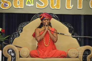 A seated man in orange-red ochre robes sits on an ornate beige throne with carved black wooden armrests and backrest. He wears a multicoloured red, pink and purple turban with a red bindi on his forehead, and a long golden bead necklace of ceremonial mala beads. He presses his palms together in anjali namaskar with eyes closed, in front of a microphone on a stand. Behind him hang navy blue drapes, and a floral arrangement of red roses and greenery sits at the left edge. Above is a gold-lettered banner partly reading Sutra.