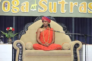 A man in an orange saffron robe with a red and purple turban sits cross-legged on a tan upholstered throne with carved black wooden arms and back. He wears a long multi-strand gold bead necklace and holds his hands folded in his lap while speaking into a microphone on a stand. Behind him hangs a green banner with gold glitter letters reading Yoga Sutras. A vase of red and white flowers stands at the left edge of a stage draped with navy curtains. Swamiji (probable): the seated figure wears a red and purple turban, orange robes and a gold bead necklace on a throne, consistent with the page's account of a talk.