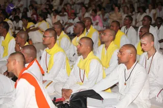 A large seated audience fills a hall, mostly men in front wearing white kurta-style clothing with bright yellow shawls draped over their shoulders. Several have shaved heads, rudraksha malas and white forehead markings, and sit with hands folded or eyes closed. Behind them, women in colourful saris sit among the crowd. A small pile of books and a black bag lie on the floor in the foreground, and a microphone is visible at lower left. No speaker's dais or a person figure is clearly visible in this frame.