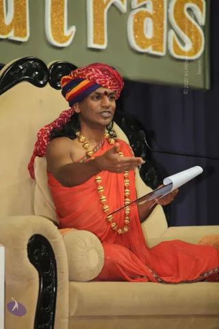 A man seated on an ornate cream upholstered throne with carved black wooden armrests speaks while gesturing with his right hand and holding a rolled paper in his left. He wears a multicoloured red, orange, and purple bandhani turban, a red dot on his forehead, an orange-red shawl over bare shoulders, and a heavy gold bead necklace. A microphone stand is visible at right. Behind him a green banner displays glittering gold letters reading 'utras' from a larger word, matching the Yoga Sutras session. Swamiji (probable): the seated speaker has a distinctive saffron-orange robe, ornate gold beaded mala and a colourful turban, consistent with the page's Swamiji reference and the in-person talk setting.