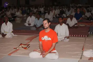 A photograph of a large indoor meditation hall with many people seated cross-legged on thin mats in rows. In the foreground a man with short dark hair and a trimmed beard sits with eyes closed, hands folded in his lap, wearing an orange T-shirt printed with a white CRUSH 2005 logo over white loose trousers. Behind him others in white ritual clothes sit in meditation, and a folded orange and white cloth lies on the mat at left.