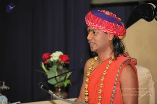 A man speaks at a microphone on a stand in front of a dark navy curtain, with a vase of red and white roses to his left and a cream upholstered chair behind him. He wears a red and purple tie-dye turban with a small red tilak on his forehead, long dark hair falling to his shoulders, and an orange-red saffron-style shawl with a decorative border. A long necklace of gold beads hangs across his chest. He appears to be giving a talk. Swamiji (probable): long swept-back hair, a colourful turban, saffron clothing and a gold bead mala identify him as the speaker, matching the page's account of a Nithyananda Satsang talk.