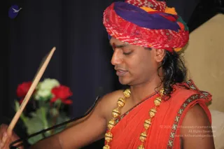 A man in a red and purple bandhani-patterned turban with a small red tilak on his forehead sits before a microphone, eyes lowered, holding a wooden stick or fan in one hand. He wears a saffron-orange garment with green and gold border trim and a long gold bead necklace. Behind him are red and white roses on a dark blue curtain.